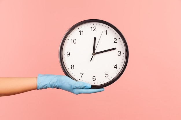 Vue latérale du profil gros plan de la main humaine dans des gants chirurgicaux bleus tenant une horloge analogique.