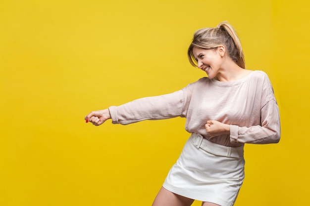 Vue latérale du profil d'une jeune femme affirmée positive aux cheveux blonds en chemisier beige décontracté debout avec un geste de traction beaucoup d'effort face à l'expression studio d'intérieur tourné isolé sur fond jaune