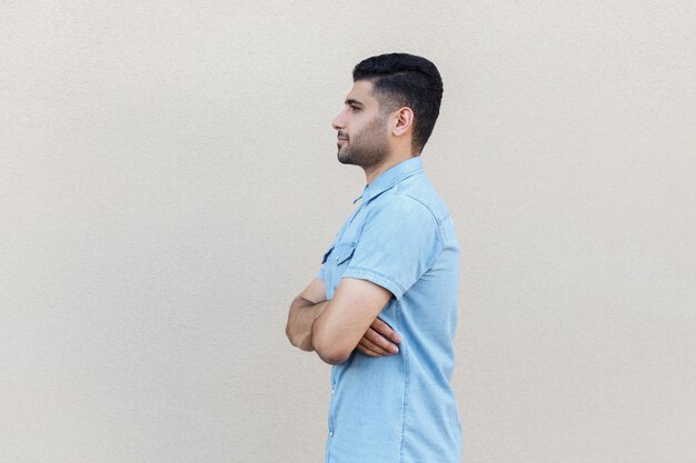 Vue latérale du profil portrait d'un beau jeune homme barbu calme et sérieux en chemise bleue debout, bras croisés et impatient. studio intérieur tourné isolé sur fond de mur beige clair.