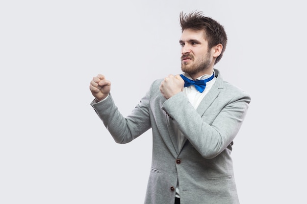 Vue latérale du profil portrait d'un bel homme barbu sérieux en costume gris décontracté et noeud papillon bleu debout avec des poings de boxe et regardant droit. tourné en studio intérieur, isolé sur fond gris clair.