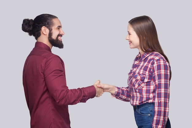 Vue latérale du profil portrait d'un homme barbu heureux avec des cheveux noirs et une femme dans un style décontracté debout et se serrant la main avec un sourire à pleines dents. tourné en studio intérieur, isolé sur fond gris.