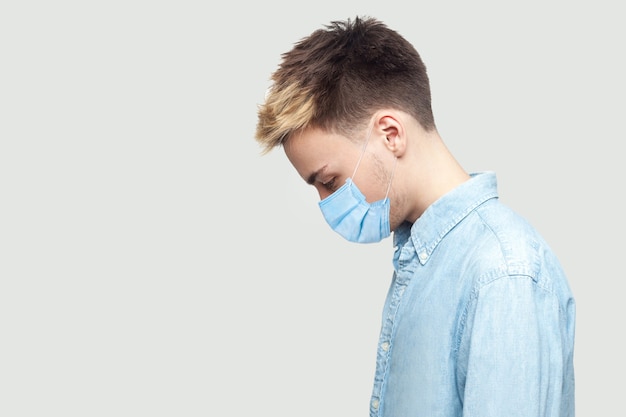 Vue latérale du profil portrait d'un jeune homme triste et seul avec un masque médical chirurgical en chemise bleue debout, tenant la tête baissée et déprimé. studio intérieur tourné sur fond gris espace copie.