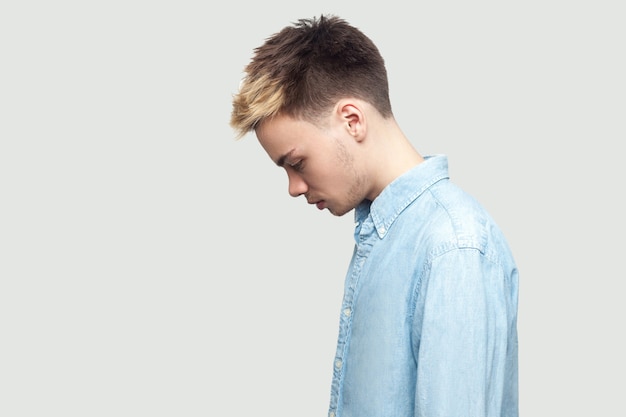 Vue latérale du profil portrait de triste bouleversé seul beau jeune homme en chemise bleu clair debout, tenant la tête baissée et déprimé. studio intérieur tourné sur fond gris espace copie.