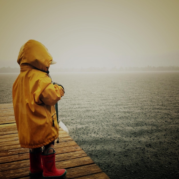 Photo vue latérale d'un enfant en manteau pendant la pluie alors qu'il se tient sur la jetée contre le ciel.