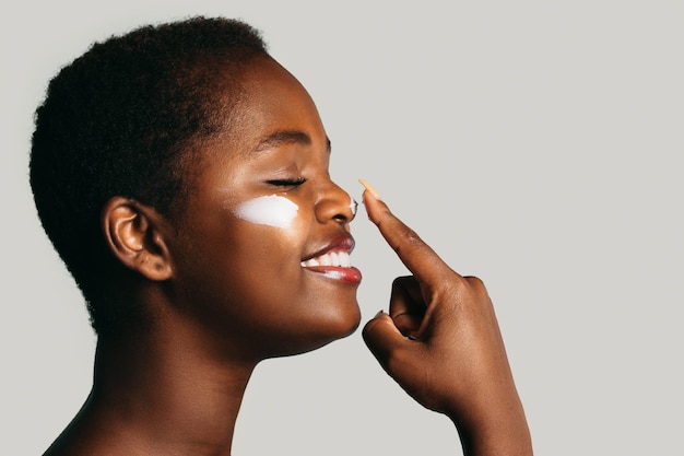 Vue latérale d'une femme afro-américaine souriante applique de la crème sur le visage isolé sur fond blanc avec ...