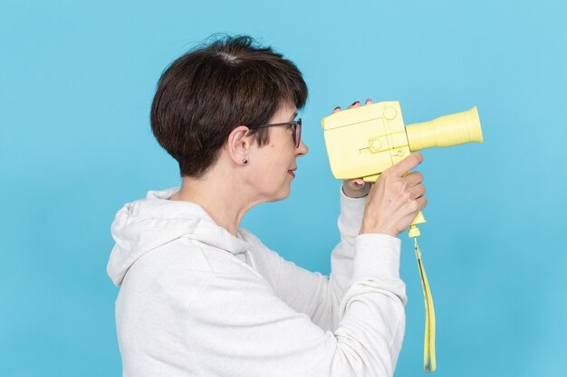 Vue latérale d'une femme d'âge moyen avec une coupe courte dans un pull et des lunettes tenant une caméra jaune vintage. Concept de tournage vidéo. espace promo