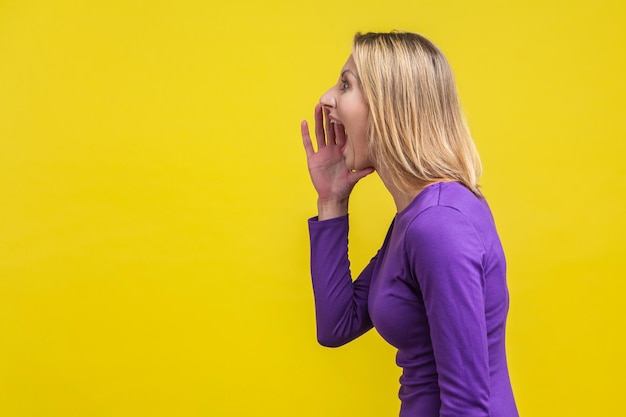 Vue latérale d'une femme agressive en robe violette élégante tenant ses mains près de la bouche grande ouverte et criant annonce, visage furieux fou. studio d'intérieur tourné isolé sur fond jaune