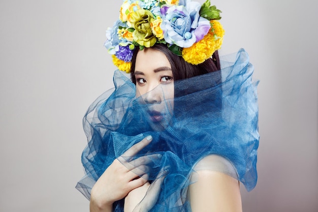 Vue latérale d'une femme asiatique sensuelle au chapeau floral avec voile bleu sur le cou posant sur fond gris clair. tourné en studio intérieur.