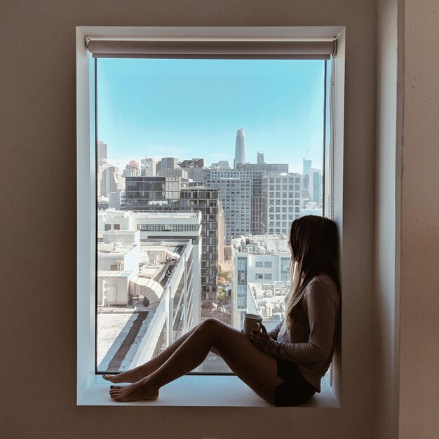 Photo vue latérale d'une femme assise sur le seuil de la fenêtre en regardant à travers la fenêtre