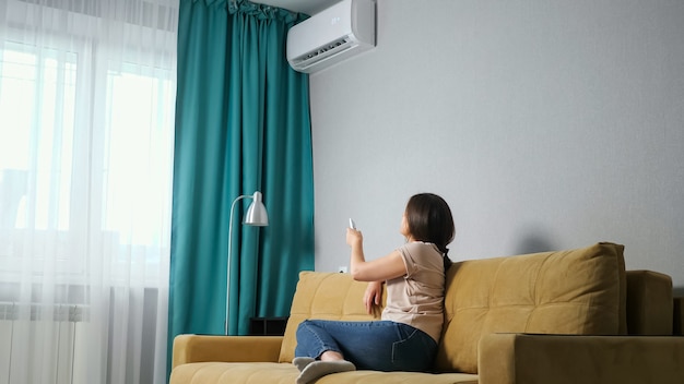 Vue latérale d'une femme brune ajuste le climatiseur assis sur le canapé.