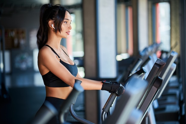 Vue latérale d'une femme brune en vêtements de sport noirs et écouteurs sans fil faisant un entraînement cardio dur sur orbitrek.