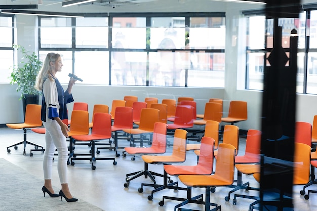 Vue latérale d'une femme caucasienne aux longs cheveux blonds, portant des vêtements élégants, debout dans une salle de réunion moderne et vide, entraînant son discours pour la conférence.