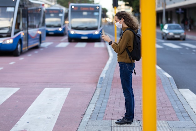 Photo vue latérale d'une femme caucasienne dans les rues de la ville pendant la journée, portant un masque facial contre le coronavirus covid19 debout et utilisant son smartphone avant de traverser la route