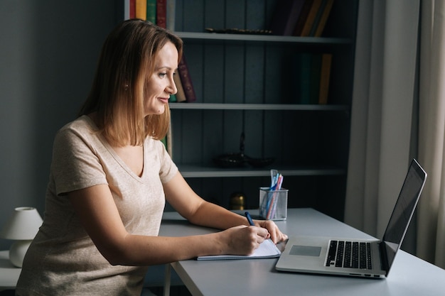 Vue latérale d'une femme concentrée écrivant en prenant des notes en étudiant en ligne sur un ordinateur dans le salon en regardant l'écran