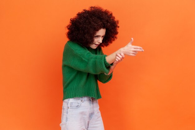 Vue latérale d'une femme dangereuse avec une coiffure afro portant un pull décontracté vert pointant un pistolet à doigt, menaçant de tuer, tirant avec un geste d'arme. Studio intérieur tourné isolé sur fond orange.