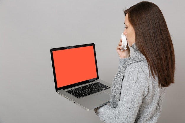 Vue latérale d'une femme en écharpe avec une serviette en papier tenant un ordinateur portable avec un écran vide vierge isolé sur fond gris. Traitement en ligne pour un mode de vie sain consultant le concept de saison froide.