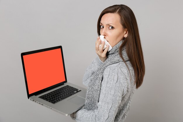 Vue latérale d'une femme en écharpe avec une serviette en papier tenant un ordinateur portable avec un écran vide vierge isolé sur fond gris. Traitement en ligne pour un mode de vie sain consultant le concept de saison froide.
