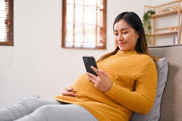 Vue latérale d'une femme enceinte asiatique heureuse utilisant son téléphone portable sur un canapé