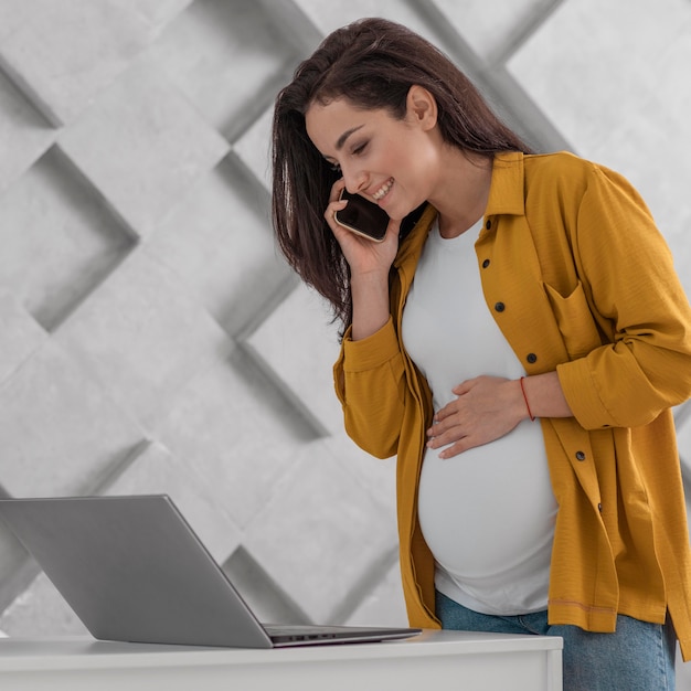 Vue latérale d'une femme enceinte travaillant avec un ordinateur portable à la maison et parler au téléphone