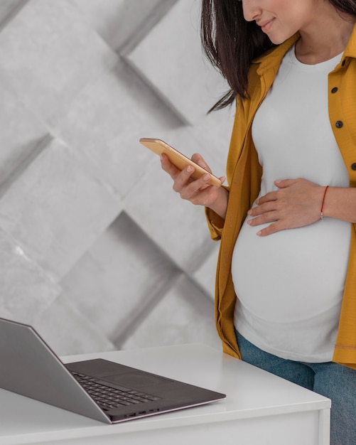 Photo vue latérale d'une femme enceinte travaillant avec un ordinateur portable et un smartphone à la maison