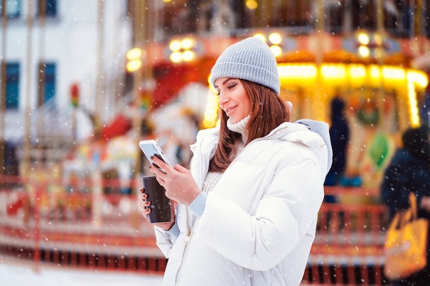 Vue Latérale D'une Femme Heureuse Portant Des Vestes Lit Un Smartphone Dans La Ville De Noël Tout En Tenant Du Café