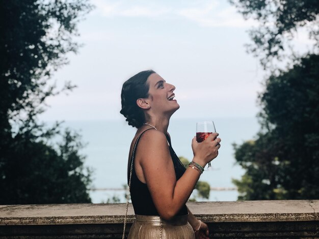 Photo vue latérale d'une femme joyeuse buvant du vin alors qu'elle se tient face à la mer