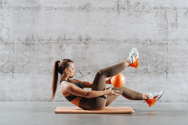 Vue latérale d'une femme puissante musclée avec queue de cheval et vêtements de sport faisant des abdos avec ballon sur le tapis en face du mur gris.