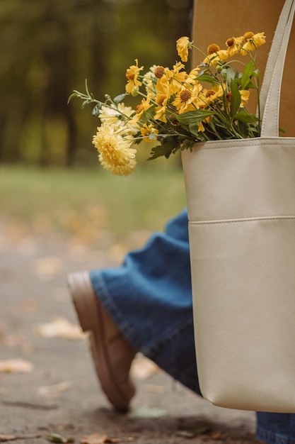 Vue latérale d'une femme de récolte tenant un sac avec un bouquet de fleurs jaunes en marchant dans le parc d'automne