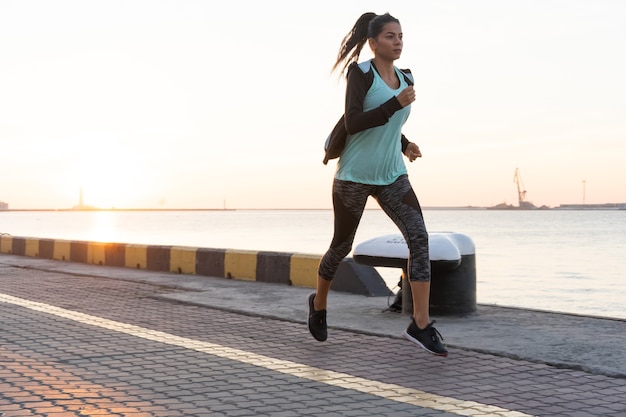 Vue latérale d'une femme de remise en forme qui court sur une route au bord de la mer près du port maritime. Sportive s'entraînant sur la promenade du bord de mer au coucher du soleil.