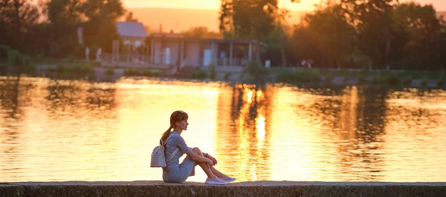 Vue latérale d'une femme seule assise seule au bord du lac par une chaude soirée. Solitude et détente dans le concept de la nature.