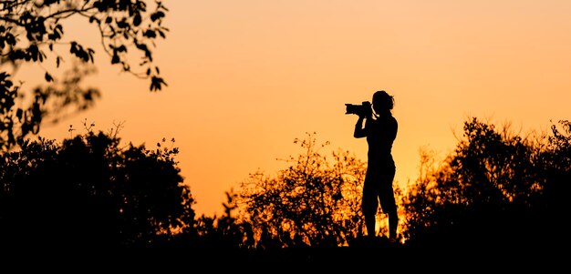 Photo vue latérale d'une femme en silhouette photographiant à l'aide d'un appareil photo numérique alors qu'elle se tient près des plantes contre un ciel clair au coucher du soleil