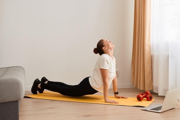 Vue latérale d'une femme en t-shirt et leggins pratiquant le yoga à la maison tout en regardant une vidéo en ligne de remise en forme sur un ordinateur portable étirant son corps à la recherche d'un mode de vie sain