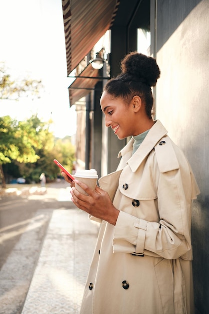 Vue latérale d'une fille afro-américaine décontractée souriante en trench-coat élégant avec du café pour aller joyeusement en utilisant un téléphone portable en plein air