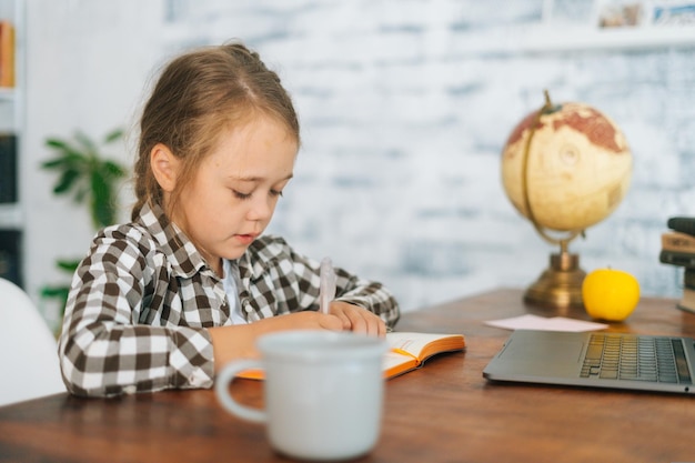 Vue latérale d'une fille de l'école primaire ciblée faisant des devoirs d'écriture assis à table avec un ordinateur portable