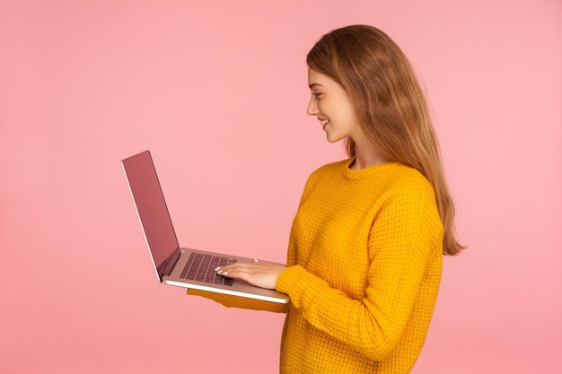 Vue latérale d'une fille gingembre souriante positive en chandail tenant un ordinateur portable, tapant sur le clavier, surfant sur Internet, étudiant ou pigiste travaillant à l'aide d'un ordinateur. studio d'intérieur tourné isolé sur fond rose