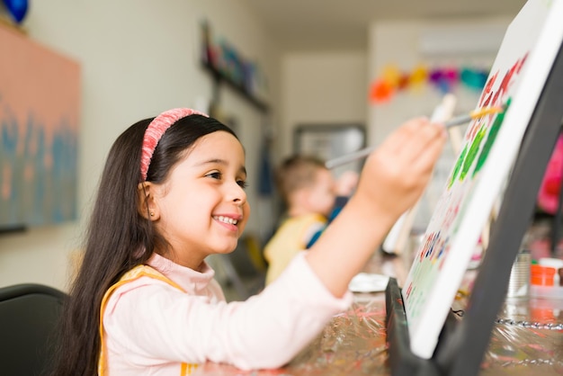 Vue latérale d'une fillette latine heureuse peignant un paysage coloré avec un pinceau sur une toile pendant son cours d'art pour enfants