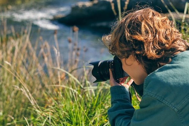 Photo vue latérale d'un garçon anonyme prenant une photo avec un appareil photo sur la côte herbeuse près de l'eau qui coule dans la nature sur un fond flou