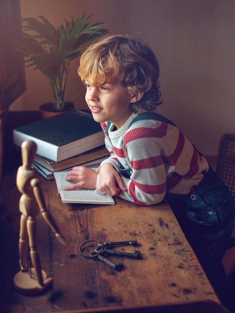 Vue latérale d'un garçon aux cheveux lus regardant ailleurs pensivement tout en étant assis à une vieille table avec un livre ouvert