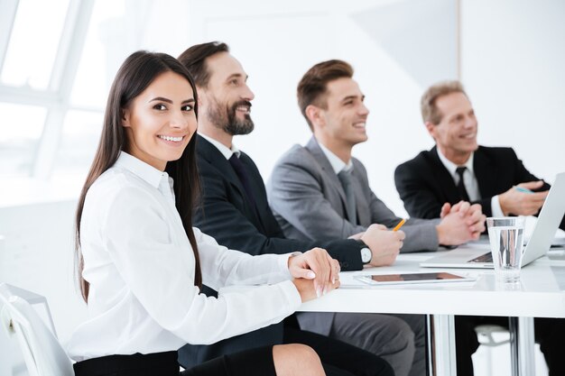 Vue latérale des gens d'affaires souriants assis près de la table et femme regardant la caméra