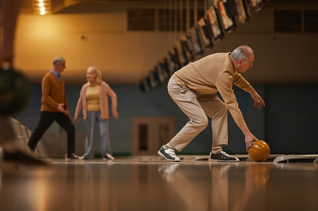 Vue latérale grand angle sur un homme âgé jouant au bowling tout en profitant d'un divertissement actif au bowling, espace pour copie