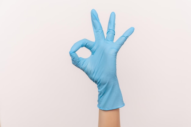 Vue latérale en gros plan de la main humaine dans des gants chirurgicaux bleus montrant le signe Ok ou le numéro 3 avec les doigts.