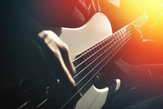 Vue latérale sur la guitare basse à cinq cordes et les mains du musicien éclairées par contraste de lumière orange dans une pièce sombre