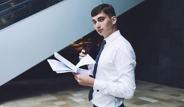 Vue latérale d'un homme d'affaires concentré dans des vêtements formels avec un stylo tenant une pile de documents dans les mains debout dans un immeuble de bureaux regardant la caméra