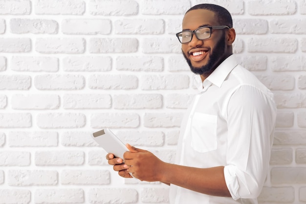 Vue latérale d'un homme afro-américain en chemise classique et lunettes à l'aide d'une tablette regardant la caméra et souriant debout contre un mur de briques blanches