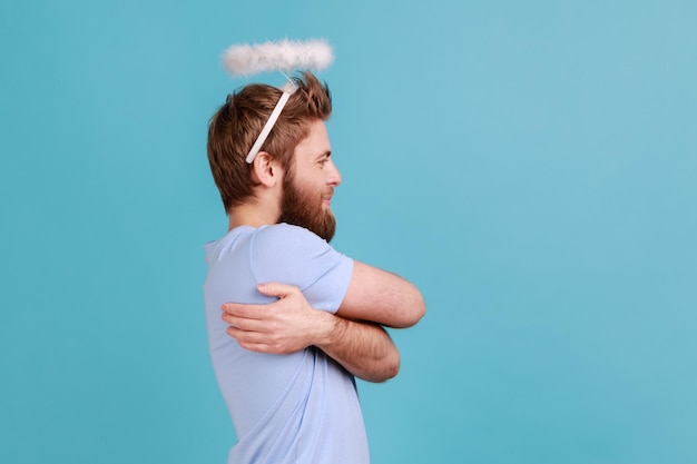 Vue latérale d'un homme angélique avec un saint nimbe debout souriant avec contentement et se serrant dans ses bras