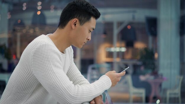 Vue latérale d'un homme asiatique hamdsome faisant défiler le téléphone sur fond de grand centre commercial