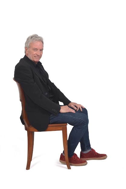 Vue latérale d'un homme assis sur une chaise avec des baskets jeans et blazer regardant la caméra sur fond blanc