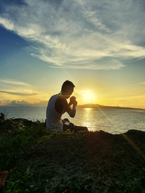 Vue latérale d'un homme assis sur un rocher sur la plage contre le ciel au coucher du soleil