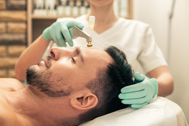 Vue latérale de l'homme aux cheveux noirs aux yeux fermés et cosmétologue dans des gants en caoutchouc touchant sa tête pendant l'ascenseur à oxygène frais