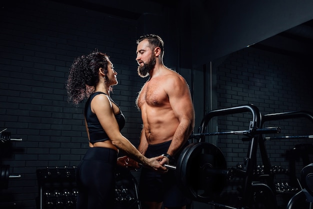 Vue latérale de l'homme barbu musclé et belle femme regardant les uns les autres et soulevant des haltères lourdes ensemble tout en exerçant dans une salle de sport sombre